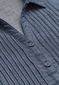 Blue, textured shirt featuring vertical pleats, a v-neckline, and dark blue buttons. Made of lightweight fabric, highlighting simplicity in design.