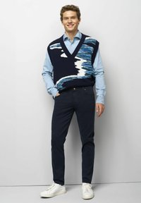 Jeune homme souriant, portant un gilet sans manches à motifs bleus sur une chemise bleu clair, un pantalon foncé et des baskets blanches, se tenant contre un mur blanc.