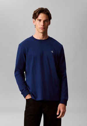 Giovane uomo che indossa una maglietta blu navy semplice a maniche lunghe con un piccolo logo bianco "cK" sul petto, posando davanti a uno sfondo grigio.