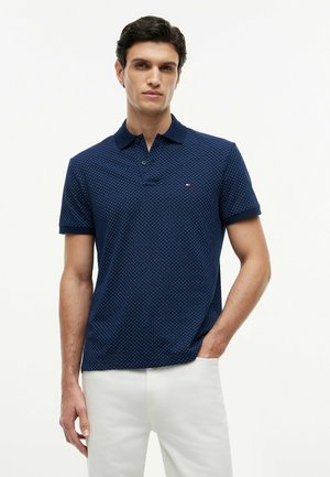 Uomo che indossa una polo blu navy con disegni a maniche corte e pantaloni bianchi, in piedi con una mano in tasca su uno sfondo semplice.