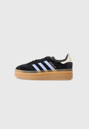 Sneaker en daim noir avec un design à trois bandes blanches, semelle en caoutchouc caramel et talon de couleur claire. Arbore le logo "Gazelle" sur le côté.
