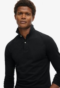 Camisa polo de manga larga negra con cuello alto y tapeta de dos botones. Fabricada con un tejido suave, presenta una textura sutil y un diseño ajustado.