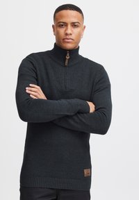 Donkergrijze gebreide trui met een hoge kraag, voorzien van een geribbeld patroon op de borst en een bruine ritssluiting. Etiketpatch nabij de zoom.