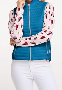 Blau gesteppte Weste mit hohem Kragen und silbernem Reißverschluss, getragen über einem pinken Oberteil mit einem Muster aus bunten Lippen. Weiße Hosen sind sichtbar.