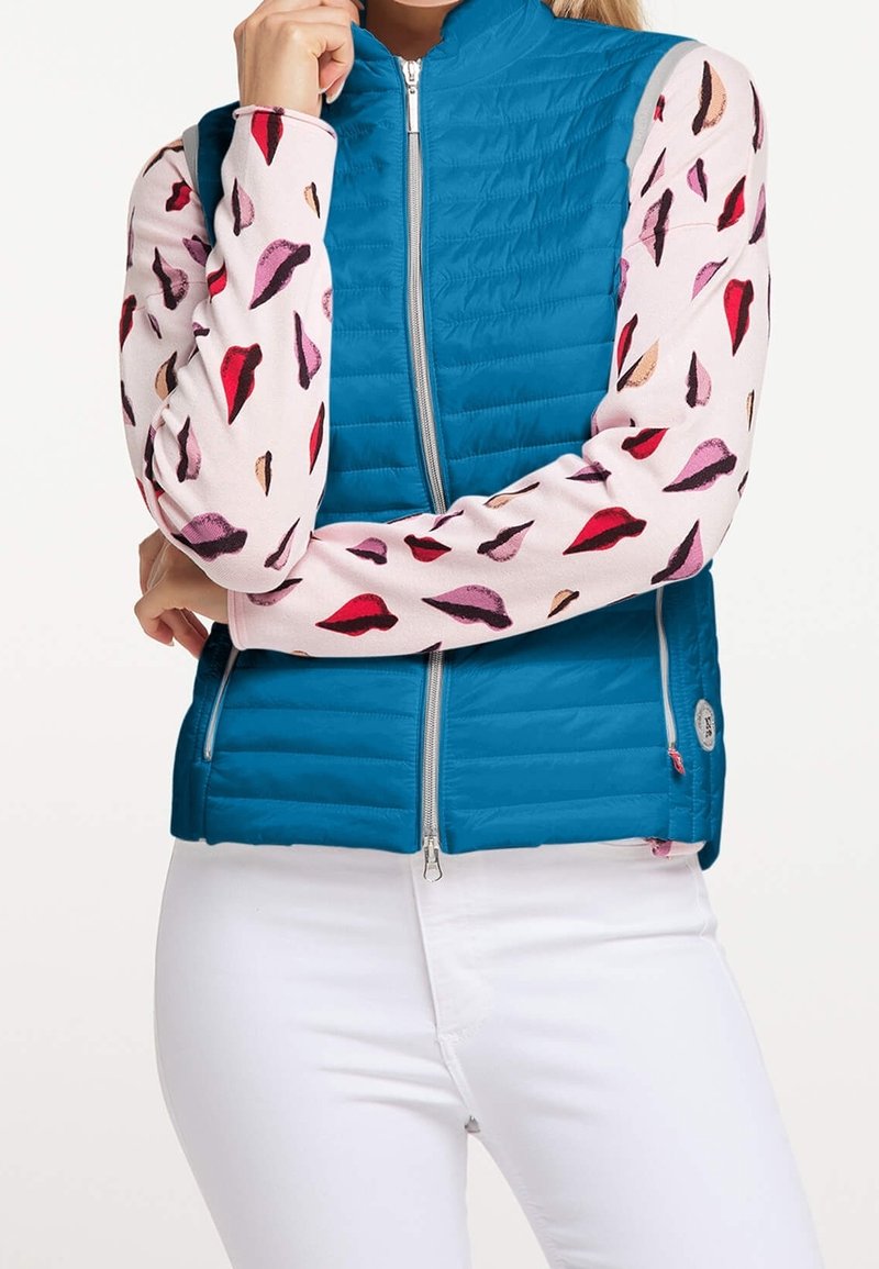 Blau gesteppte Weste mit hohem Kragen und silbernem Reißverschluss, getragen über einem pinken Oberteil mit einem Muster aus bunten Lippen. Weiße Hosen sind sichtbar.