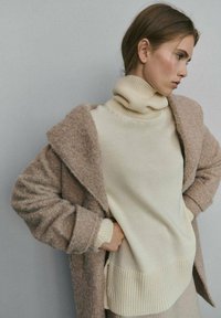 Suéter de lana beige con cuello alto y dobladillo acanalado, llevado debajo de un abrigo marrón suave y holgado con un acabado texturizado.