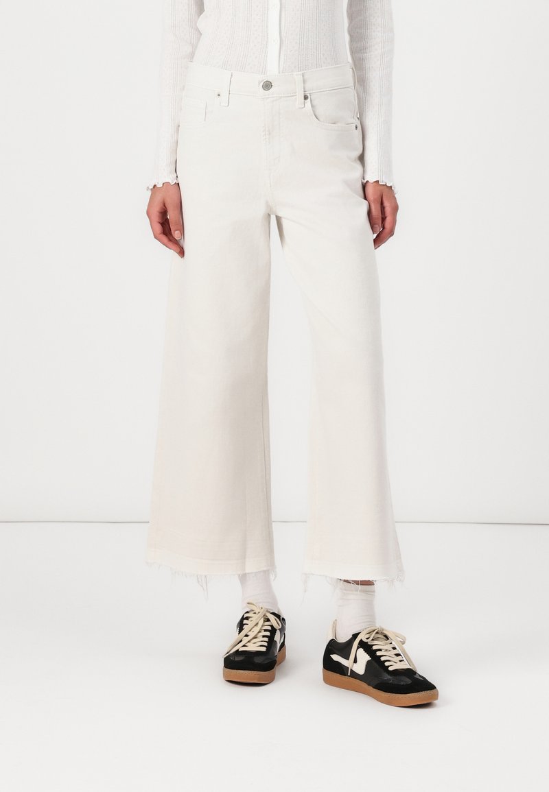 Vita vida jeans med fransad fåll, i kombination med svarta sneakers med vita accenter och ljusa strumpor. Slät denimtextur.