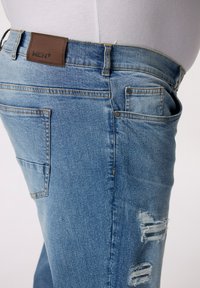 Vaqueros de mezclilla azul claro con un diseño ajustado, que presentan una etiqueta de cuero marrón, un bolsillo delantero y detalles desgastados en la zona del muslo.