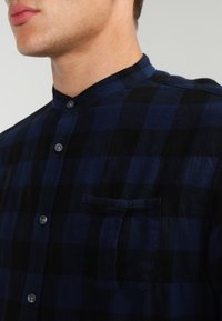 Mies, joka pukeutunut tummansiniseen ja mustaan ruudulliseen paitaan, jossa on pystykaulus ja napitettu edusta, näkyvissä rinta- ja kaula-alue.