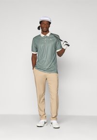 Hombre con polo a rayas verdes, pantalones beiges, gorra blanca y guante de golf, sosteniendo un palo de golf sobre el hombro, contra un fondo liso.