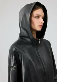 Giacca in pelle nera con cappuccio, dotata di zip frontale e tasca laterale con zip. Tessuto liscio con design aderente.