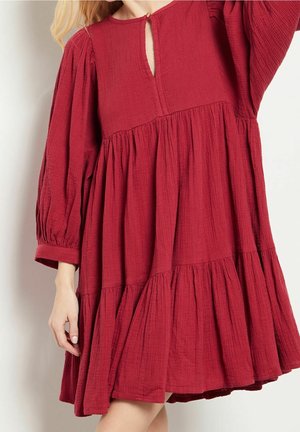 Kvinde iført en løs, langærmet, rød tekstureret kjole med opsamlede lag og en lille nøglehulshalsudskæring med knap.