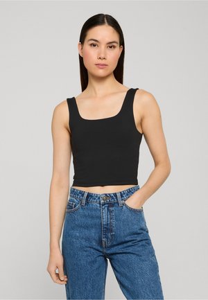 Hosszú, sötét hajú nő fekete ujjatlan crop topot és kék, magas derekú farmert visel, egy kezét zsebbe dugva áll.