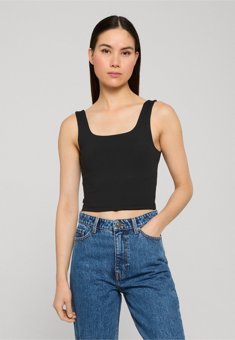 Femme aux longs cheveux foncés portant un crop top noir sans manches et un jean bleu taille haute, debout avec une main dans la poche.