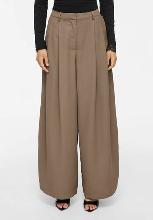 Femme portant un pantalon marron taille haute à jambes larges avec passants de ceinture, un haut noir à manches longues, et des talons ouverts noirs, debout sur fond blanc.