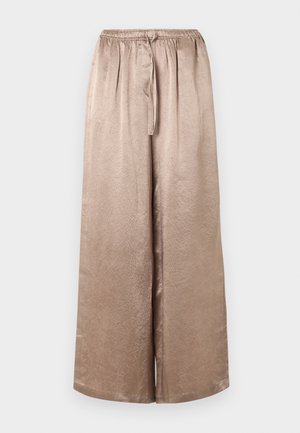 Personne grande portant une chemise ample en satin beige et un pantalon par-dessus un haut blanc avec des chaussures noires pointues, debout devant un fond uni.