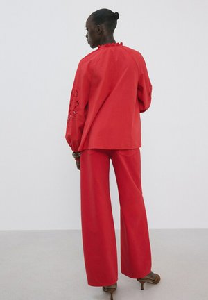 Femme à la peau foncée et aux cheveux en chignon, portant un pantalon rouge à jambes larges et un haut ample à manches longues avec des manches brodées, debout devant un fond uni.