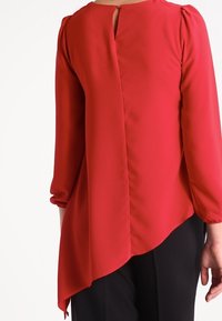 Blusa roja de manga larga con dobladillo asimétrico, que cuenta con un cierre en forma de ojo de cerradura en la parte posterior y hombros fruncidos, fabricada en una tela ligera y suave.