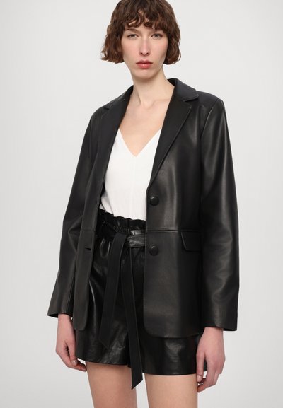 Black leather blazer with notched lapels, two front buttons, and a smooth texture, worn over a white V-neck top and black belted shorts.