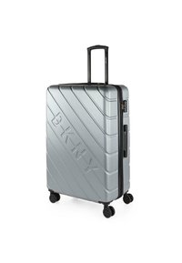 Maleta rígida plateada con surcos diagonales y logo en relieve "BKNY". Cuenta con una asa retráctil y cuatro ruedas negras para facilitar su movilidad.