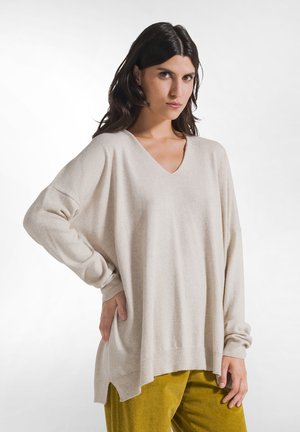 Kobieta o ciemnych włosach, ubrana w oversize beżowy sweter z dekoltem w serek oraz musztardowe spodnie, stojąca z jedną ręką na biodrze na białym tle.