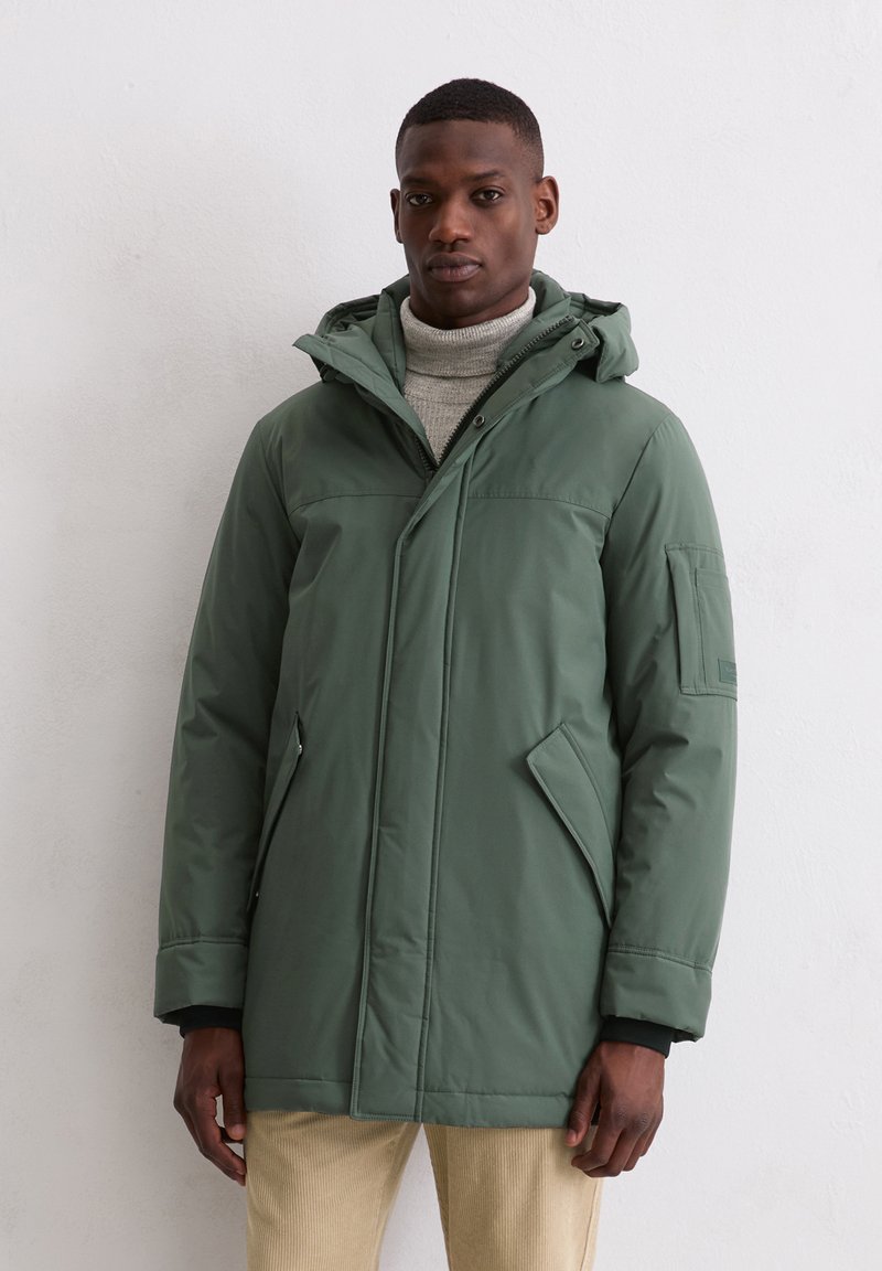 Man in een groene winterjas met capuchon, draagt een beige broek en een grijze coltrui, staand tegen een lichte, effen muur.