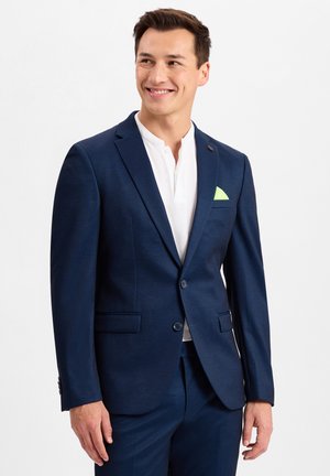 Marineblaues Sakko mit strukturiertem Finish, zwei vorderen Taschen und einem grünen Einstecktuch. Getragen über einem weißen Hemd mit Kragen.