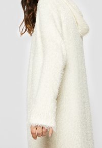 Texturerad vit cardigan med en lös passform, som har huva, långa ärmar och en fluffig tyg för ett mjukt utseende. Fluffig detalj på ärmsluten synlig.