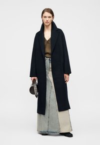 Γυναίκα που φοράει μακρύ μαύρο παλτό, μακριά φούστα από τζιν δίχρωμη, καφέ μπλούζα, μαύρα παπούτσια, κρατώντας μια στρογγυλή τσάντα με υφή.