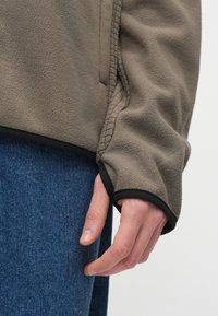 Γκρι-καφέ φλις μπουφάν με μαύρες λεπτομέρειες, διαθέτοντας ραμμένη ραφή και άνετη εφαρμογή, συνδυασμένο με μπλε τζιν παντελόνι.