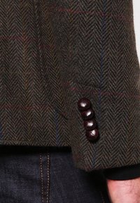 Ärmar av en brun ulljacka med fiskbensmönster som pryds av tre strukturerade vinröda knappar. Mörk denim synlig nedanför.