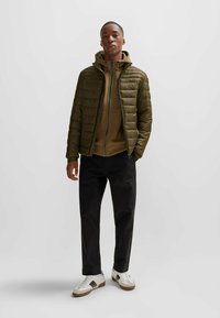 Chaqueta de abrigo color verde oliva con silueta ajustada, cierre con cremallera y capucha, superpuesta sobre una sudadera con cremallera marrón y pantalones negros. Zapatillas blancas.