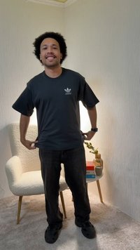 Mladý muž sa usmieva a upravuje si tmavé tričko značky Adidas v svetlej miestnosti s bielou stoličkou a malým stolíkom, na ktorom sú knihy a rastlina.