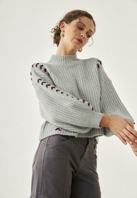 Suéter de canalé gris claro con cuello alto, mangas anchas y acentos burdeos en los hombros. Llevado con pantalones gris oscuro. Textura suave.