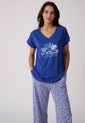 Frau trägt ein blaues V-Ausschnitt-Shirt mit maritimem Aufdruck und Text "Sea you soon,", dazu weiße und blaue Hosen mit Blattmuster, Hände in den Taschen.