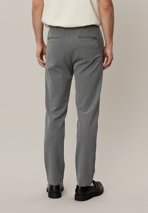 Homme portant un pantalon gris sur mesure et des mocassins noirs, debout, dos à la caméra, sur un fond uni.