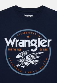 Námořnicky modré tričko s kruhovým logem, které obsahuje "Wrangler" v bílé, grafiku orla a text s oranžovými a modrými akcenty.