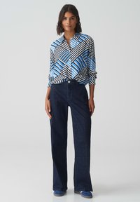 Camicia a bottoni blu e nera con motivo a righe diagonali, abbinata a jeans larghi blu scuro e scarpe blu. Tessuto dalla trama liscia.