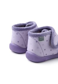 Zapatillas de tela moradas con forro suave, que presentan patrones de estrellas, correas ajustables y suelas de goma para agarre. El material es aterciopelado y texturizado.