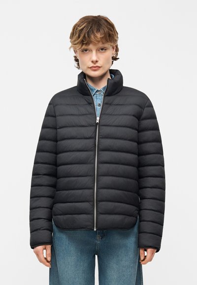 Personne portant une veste matelassée noire zippée par-dessus une chemise en jean boutonnée et un jean bleu, faisant face à l'avant sur un fond uni.