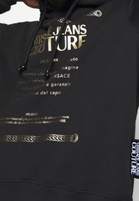 Black hoodie featuring gold text and designs, with a ribbed hem and cuffs, drawstring with black aglets, and a prominent label on the side.