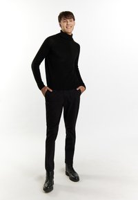 Schwarzer Rollkragenpullover mit gerippten Details, kombiniert mit schwarzen Hosen und schwarzen Schnürstiefeln, an einem Modell, das mit den Händen in den Taschen steht.