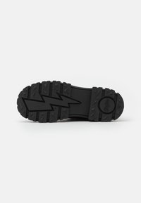 Sole di scarpa in gomma nera con scanalature profonde e un motivo a zigzag, dotata di lugs di trazione robusti e un dettaglio circolare del logo.