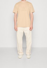 Camiseta de algodón beige con un logotipo texturizado de Adidas, combinada con pantalones sueltos color crema y zapatillas blancas. Diseño simple y minimalista.