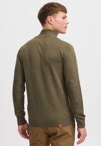 Pull en maille vert olive avec un col montant, des poignets côtelés et des coudières. Il présente une finition texturée et une petite étiquette de marque au bas.