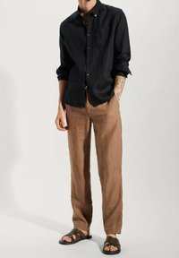 Camicia button-up nera in lino, pantaloni in lino marroni e sandali scuri. Vestibilità ampia, tessuto strutturato e design minimalista.