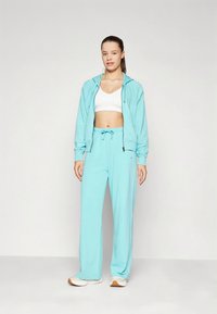 Fato de treino atlético azul claro, composto por um casaco com capuz e fecho éclair e calças de corte largo, feitos de tecido macio. É usado um sutiã desportivo branco por baixo.