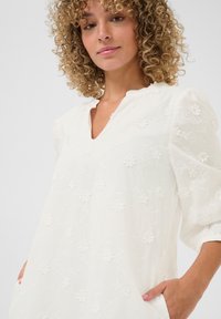 Vit broderad blus med V-ringning, blommönster, trekvartslånga puffärmar och sidfickor. Lätt och texturerad tyg.