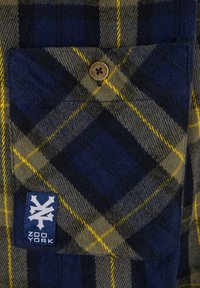 Kockás flaneling sötétkéken, szürkében és sárgában. Elől található egy mellzseb, fémgomb és egy kék logópatch, amely "ZOO YORK" felirattal látható.