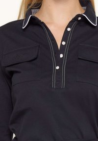 Polo shirt bleu marine avec un devant à boutons, des coutures blanches, deux poches poitrines et un col à motifs. Fabriqué en matériau en coton.